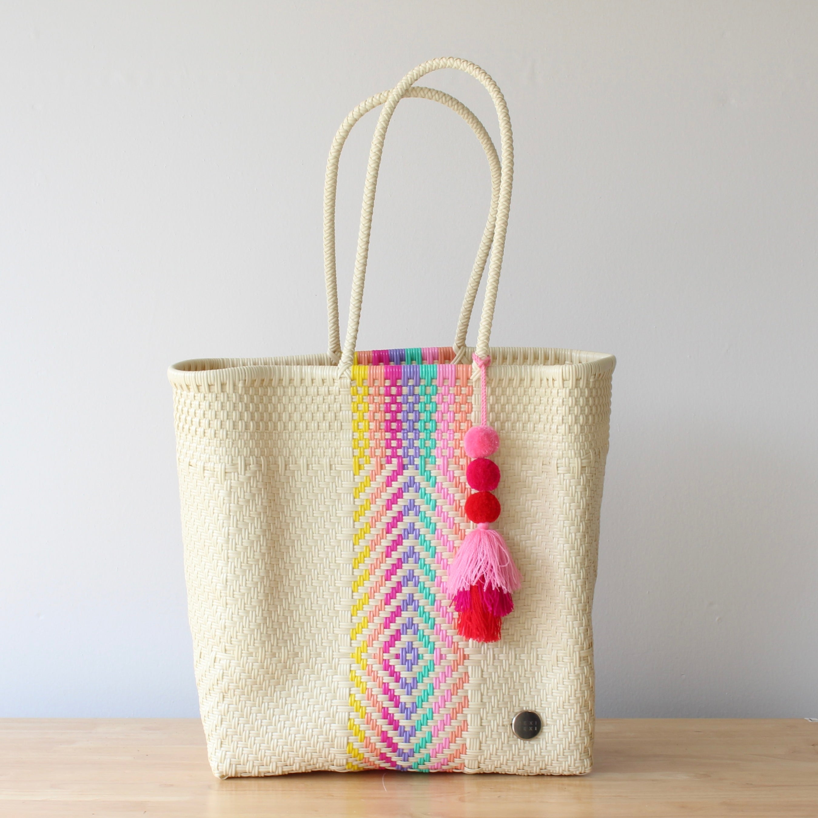 Woven tote bag with colorful striped pattern and pom-poms on a wooden surface.