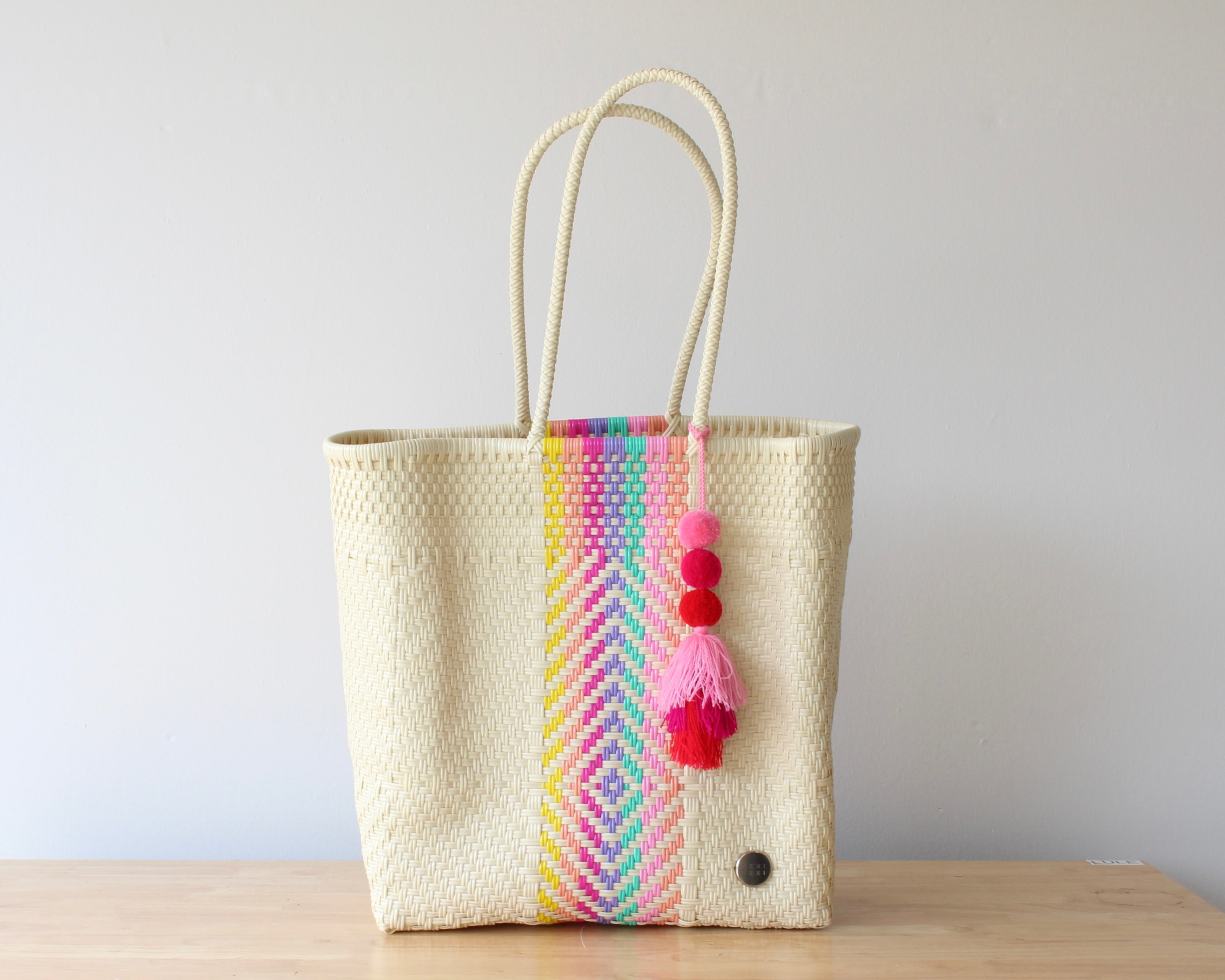 Woven tote bag with colorful striped pattern and pom-poms on a wooden surface.