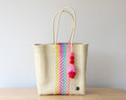 Woven tote bag with colorful striped pattern and pom-poms on a wooden surface.