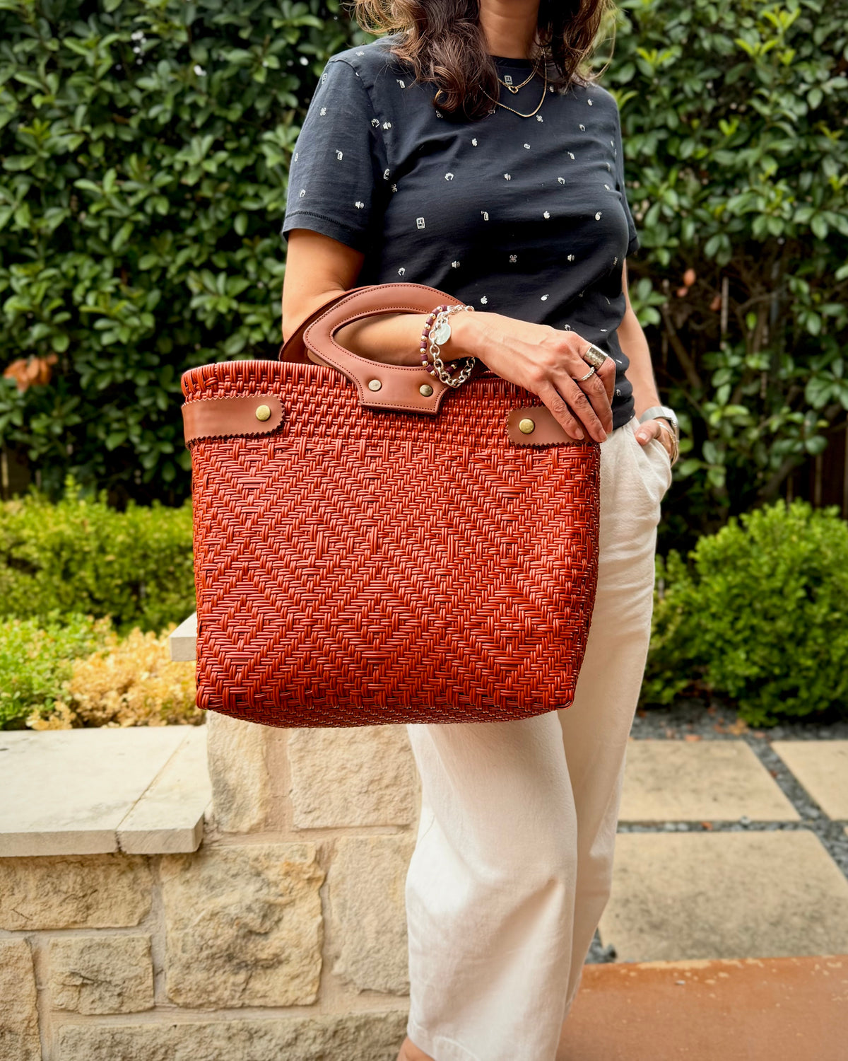 A woman wearing a black t-shirt and cream pants holds a large woven terracotta orange tote bag with brown leather handles and gold-tone hardware, set against a background of green hedges and a stone wall.