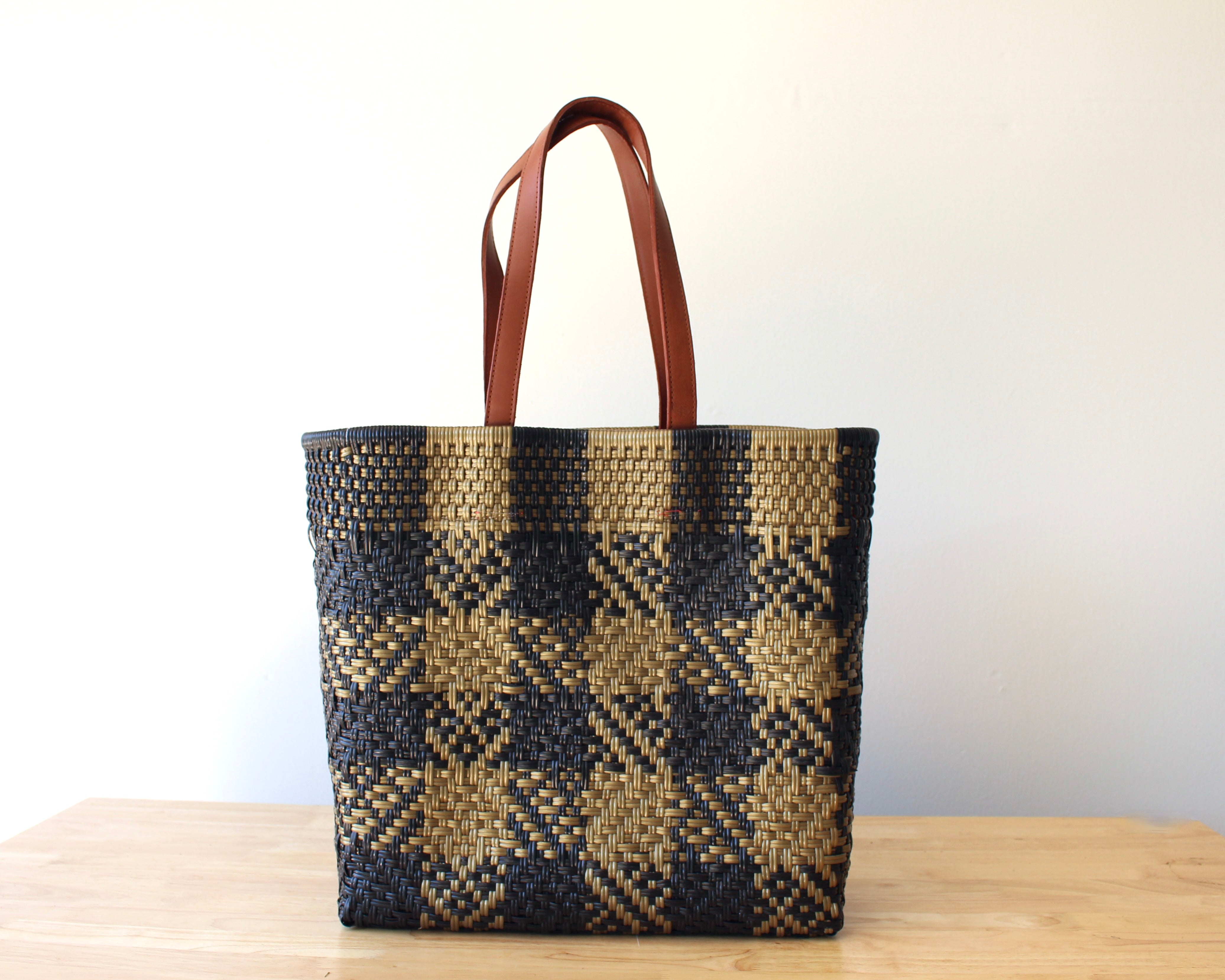 A woven tote bag with a black and tan plaid pattern and brown leather handles, standing on a light wooden surface against a plain white wall.