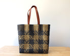 A woven tote bag with a black and tan plaid pattern and brown leather handles, standing on a light wooden surface against a plain white wall.