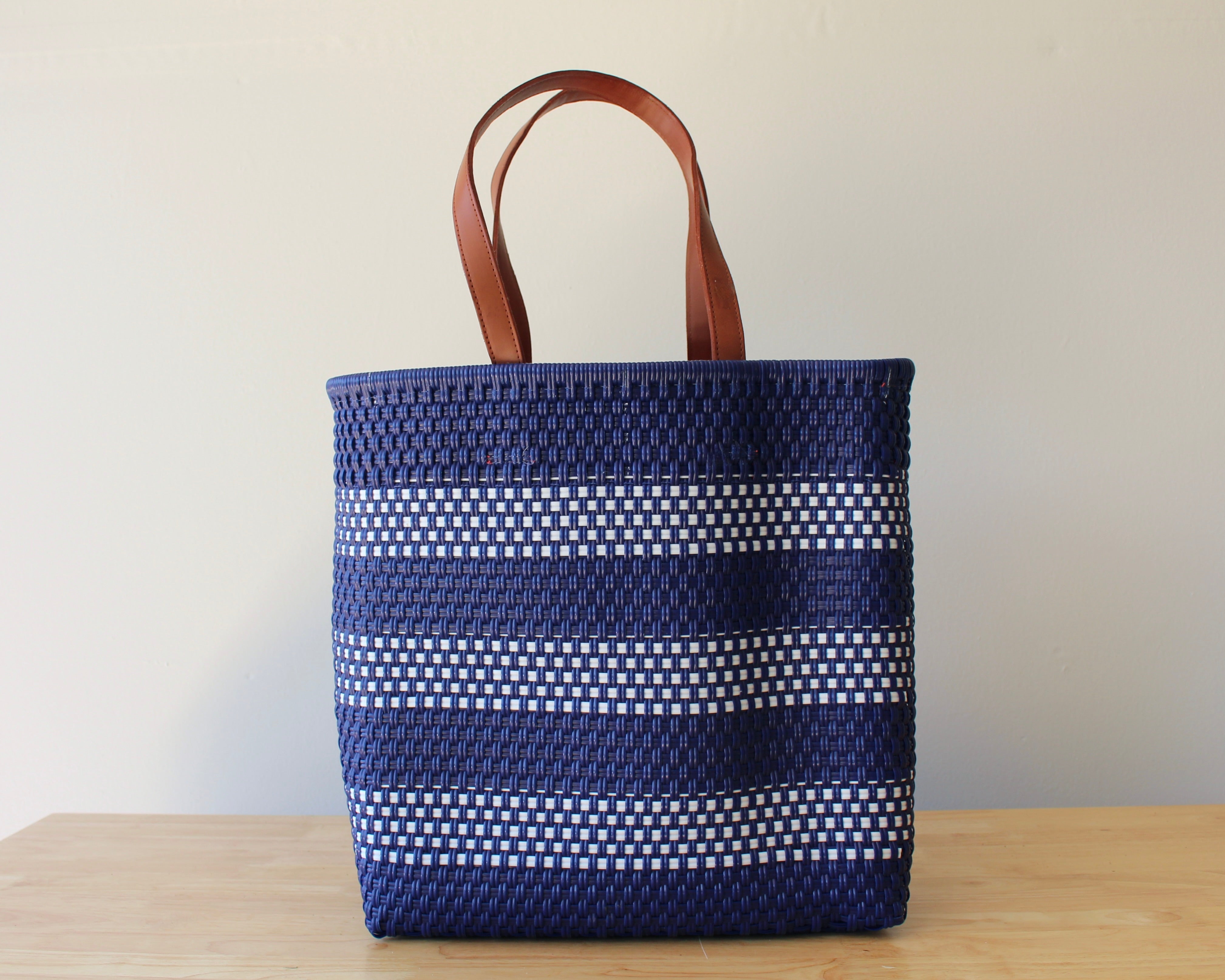 A woven tote bag with dark blue and white horizontal stripes and brown leather handles, standing on a light wooden surface against a plain beige wall.