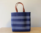 A woven tote bag with dark blue and white horizontal stripes and brown leather handles, standing on a light wooden surface against a plain beige wall.