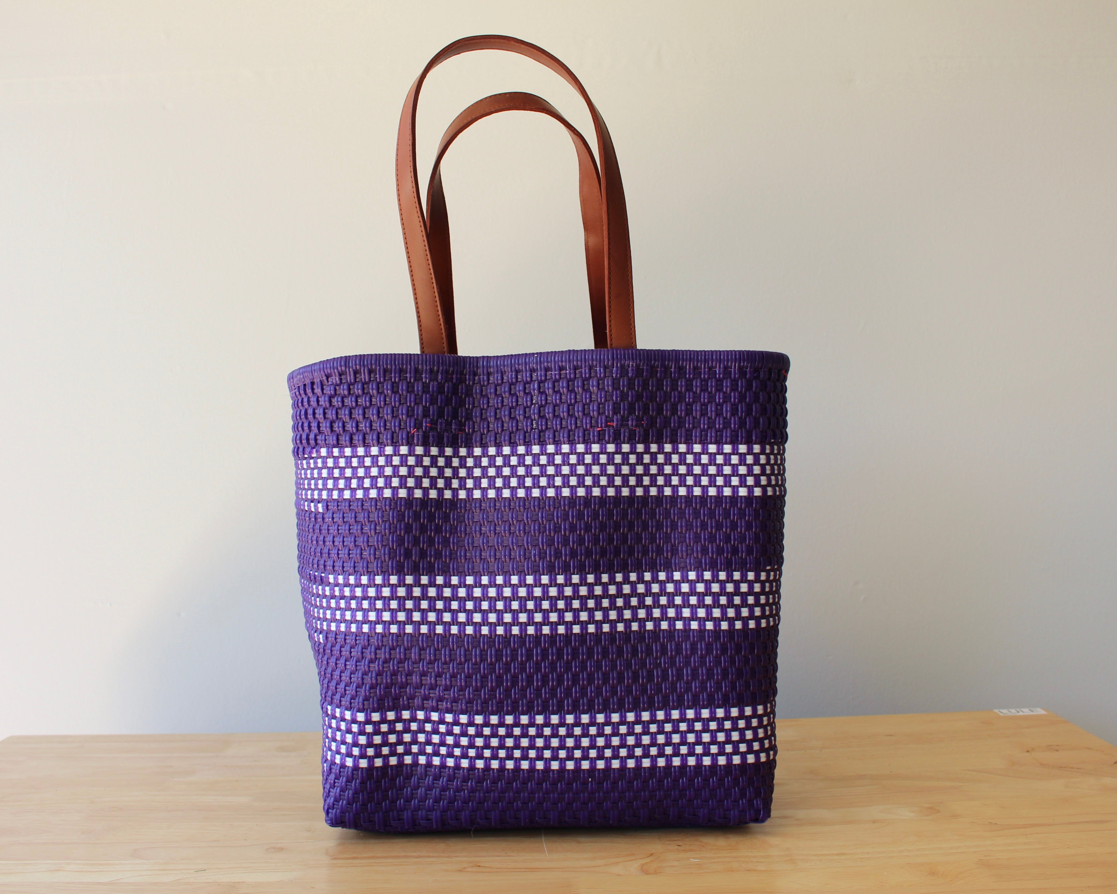 A purple woven tote bag with brown leather handles, featuring horizontal white dotted stripes, standing on a light wooden table against a plain off-white wall.