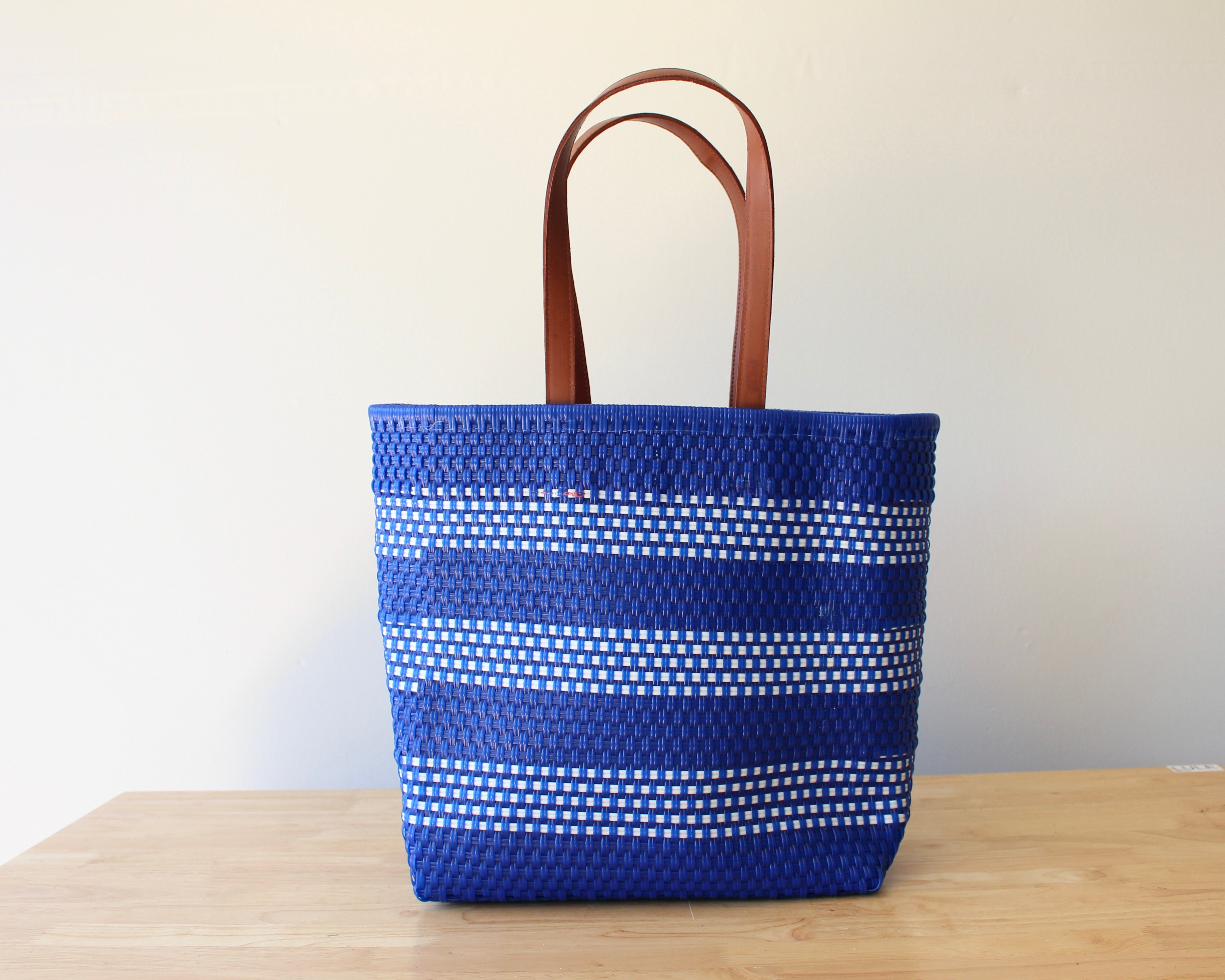 A blue and white woven tote bag with brown leather handles, standing on a light wooden table against a plain off-white wall.