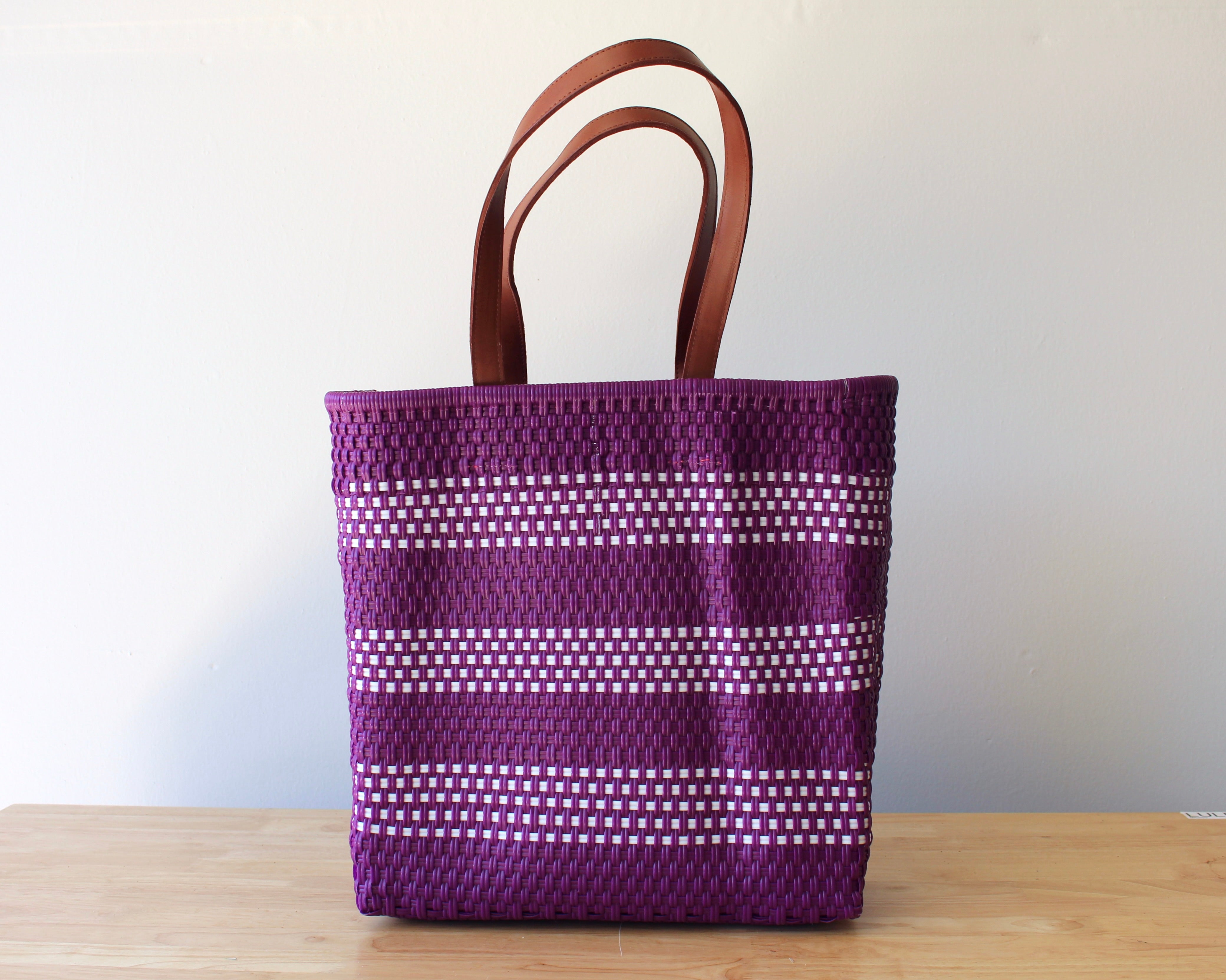 A purple woven tote bag with white dotted stripes and brown leather handles, standing on a light wooden surface against a plain off-white wall.