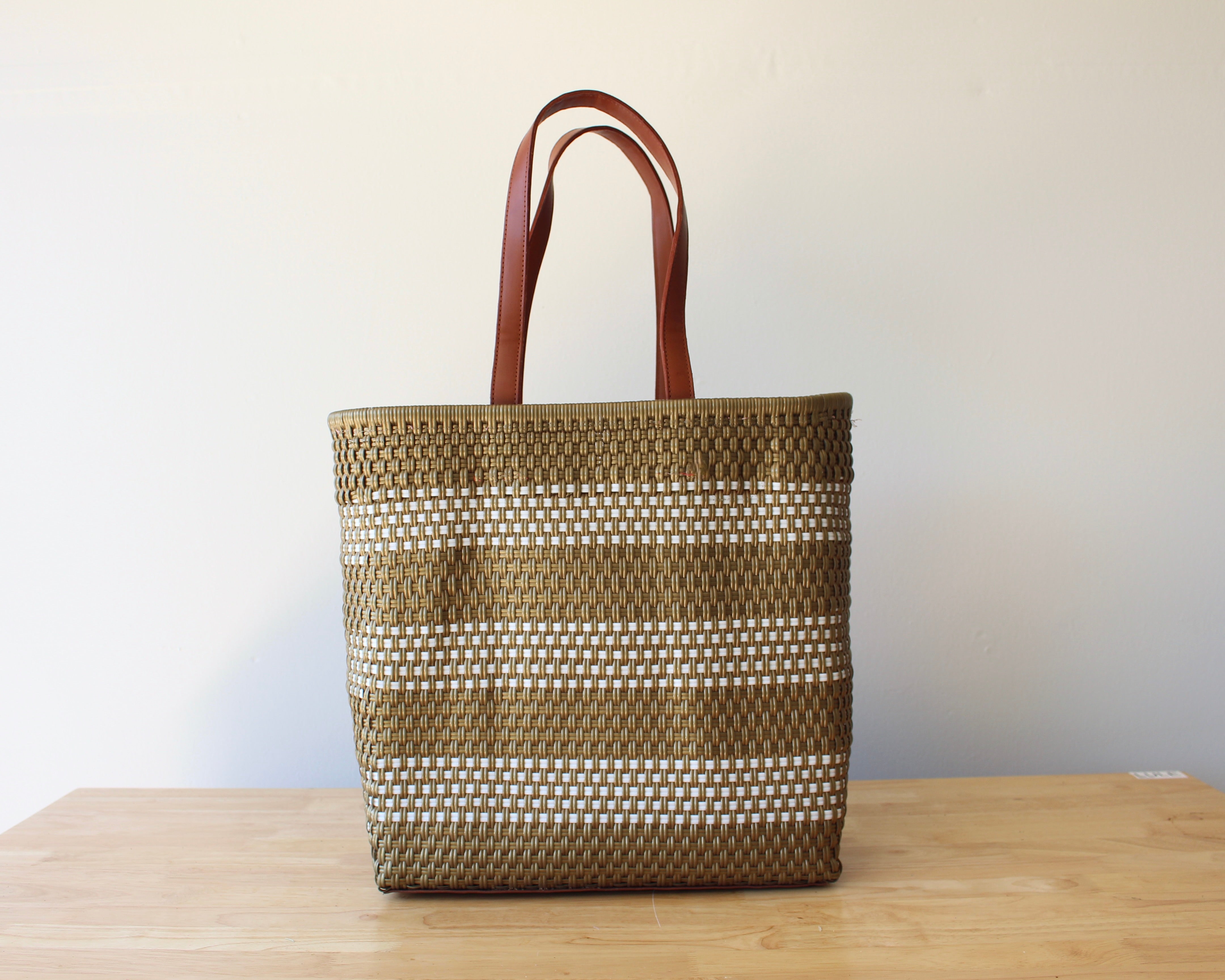 A woven tote bag with brown leather handles, featuring horizontal stripes of brown and white woven fibers, standing on a light wooden table against a plain off-white wall.