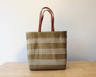 A woven tote bag with brown leather handles, featuring horizontal stripes of brown and white woven fibers, standing on a light wooden table against a plain off-white wall.