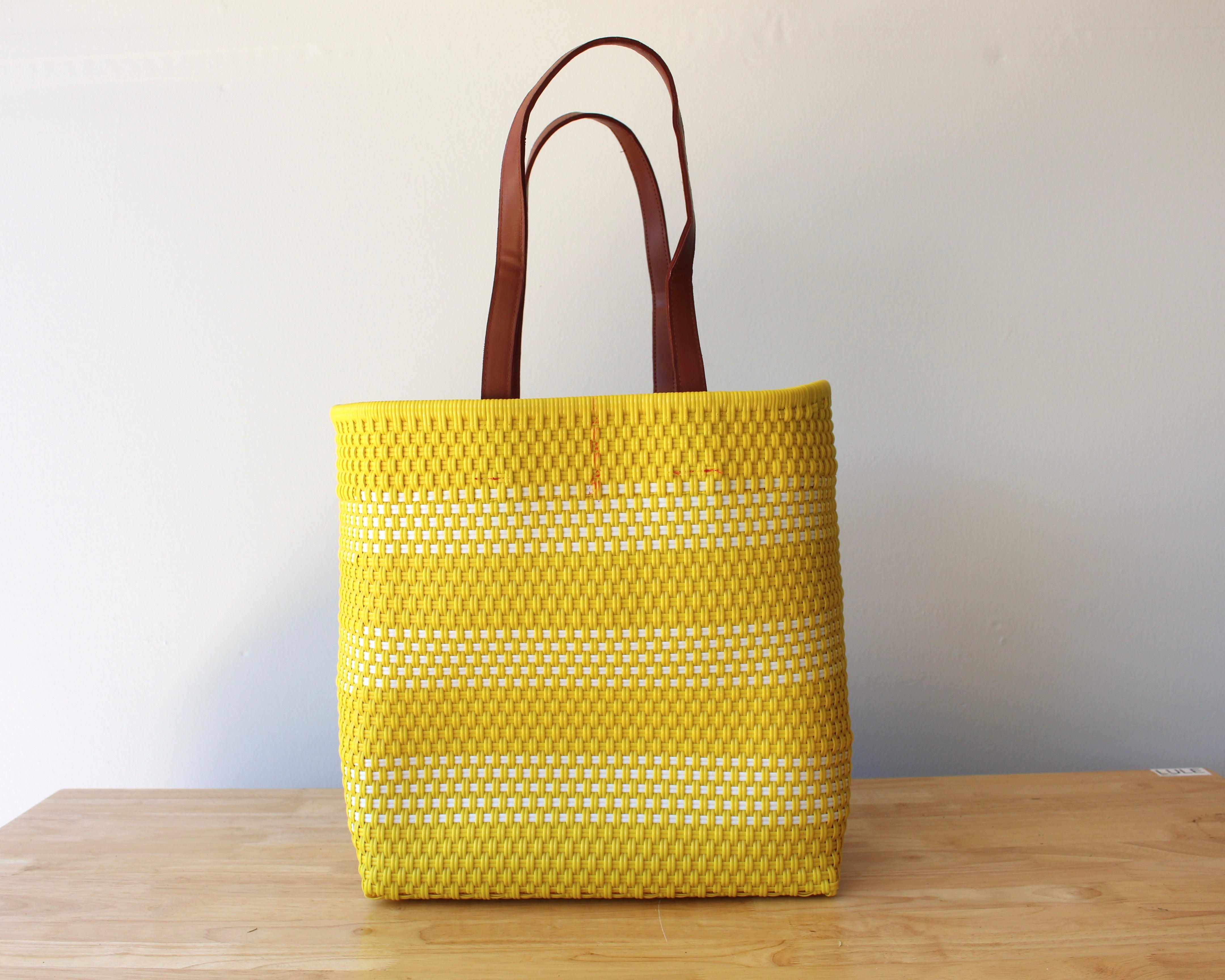 A yellow woven tote bag with brown leather handles, standing on a light wooden table against a plain off-white wall.