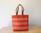 An orange woven tote bag with white dotted horizontal stripes and brown leather handles, standing on a light wood table against a plain off-white wall.