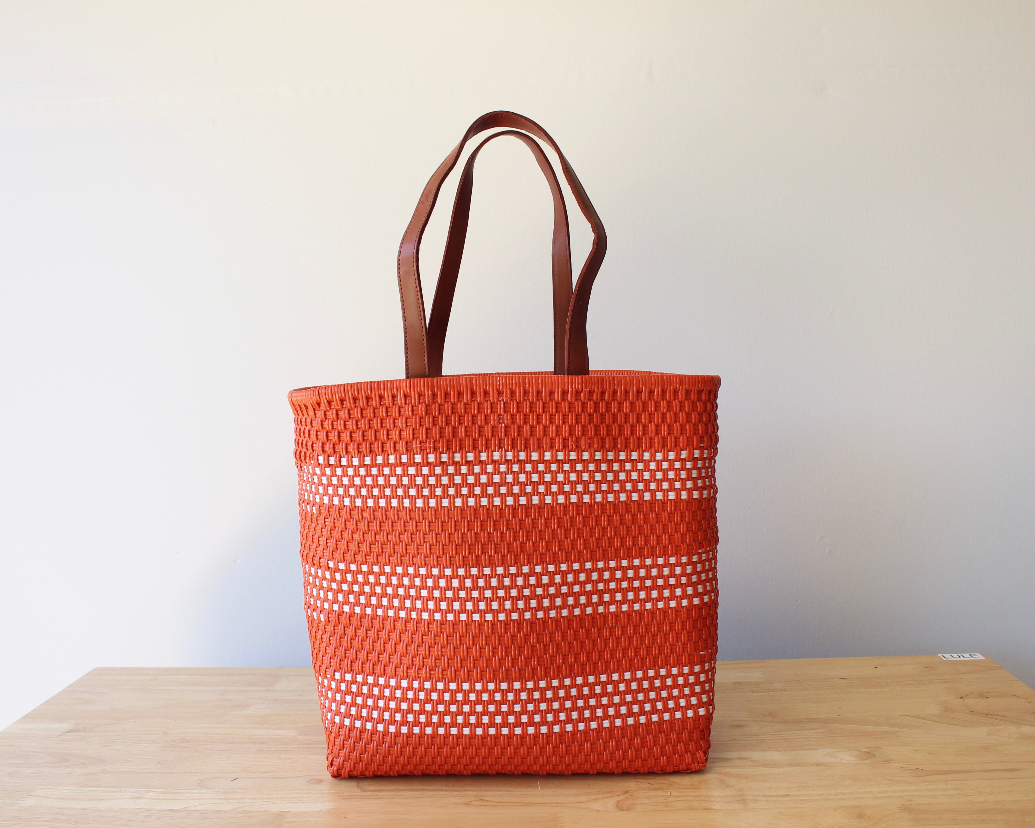 An orange woven tote bag with white dotted horizontal stripes and brown leather handles, standing on a light wood table against a plain off-white wall.