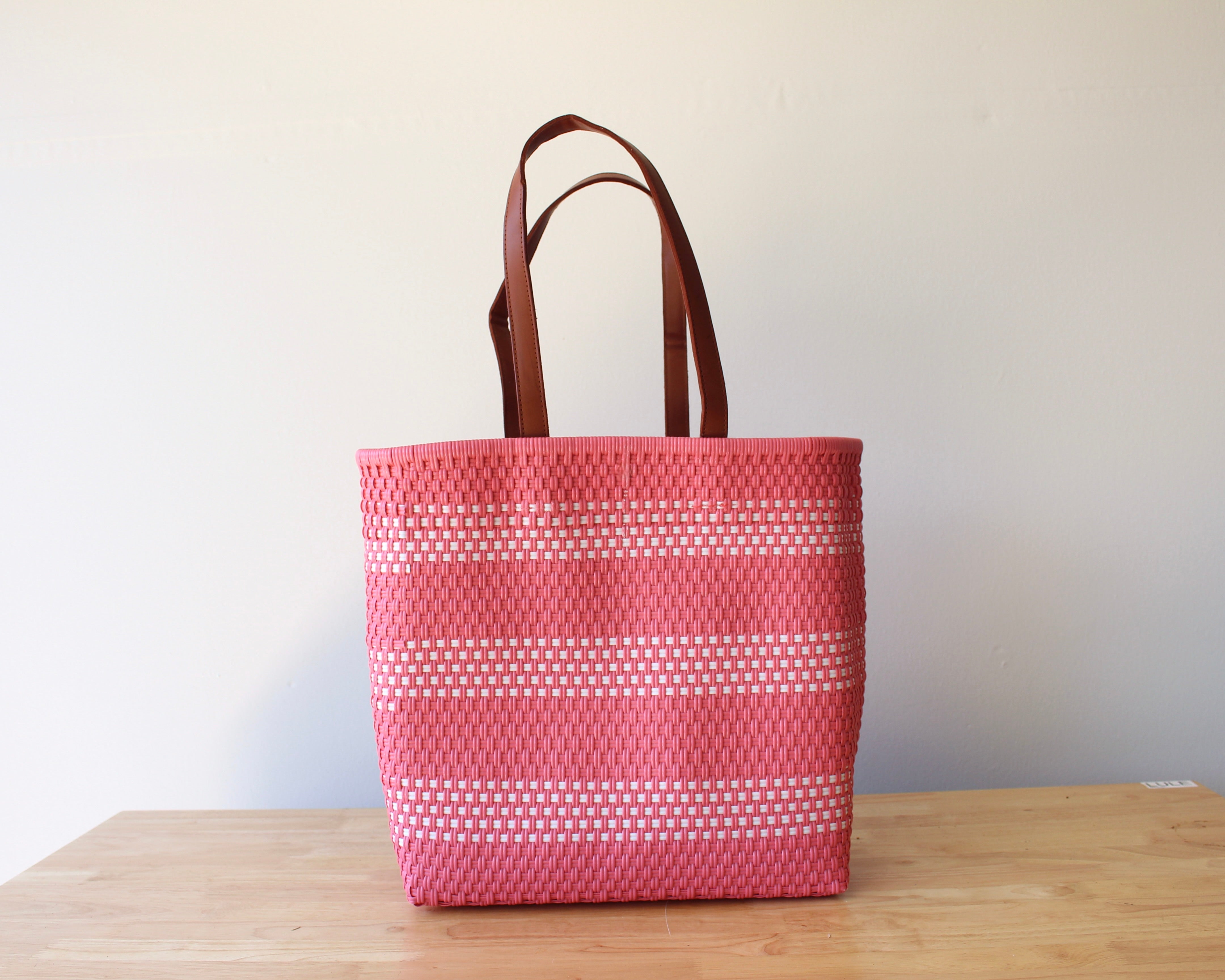 A pink woven tote bag with white dotted stripes and brown leather handles, standing on a light wooden table against a plain off-white wall.