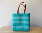 A turquoise and white woven tote bag with brown leather handles, standing on a light wooden table against a plain off-white wall.