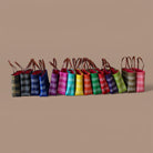 A row of Mexican woven shoulder bags in multiple colors, including black, blue, pink, yellow, green, orange, red, and brown, arranged against a solid beige background.